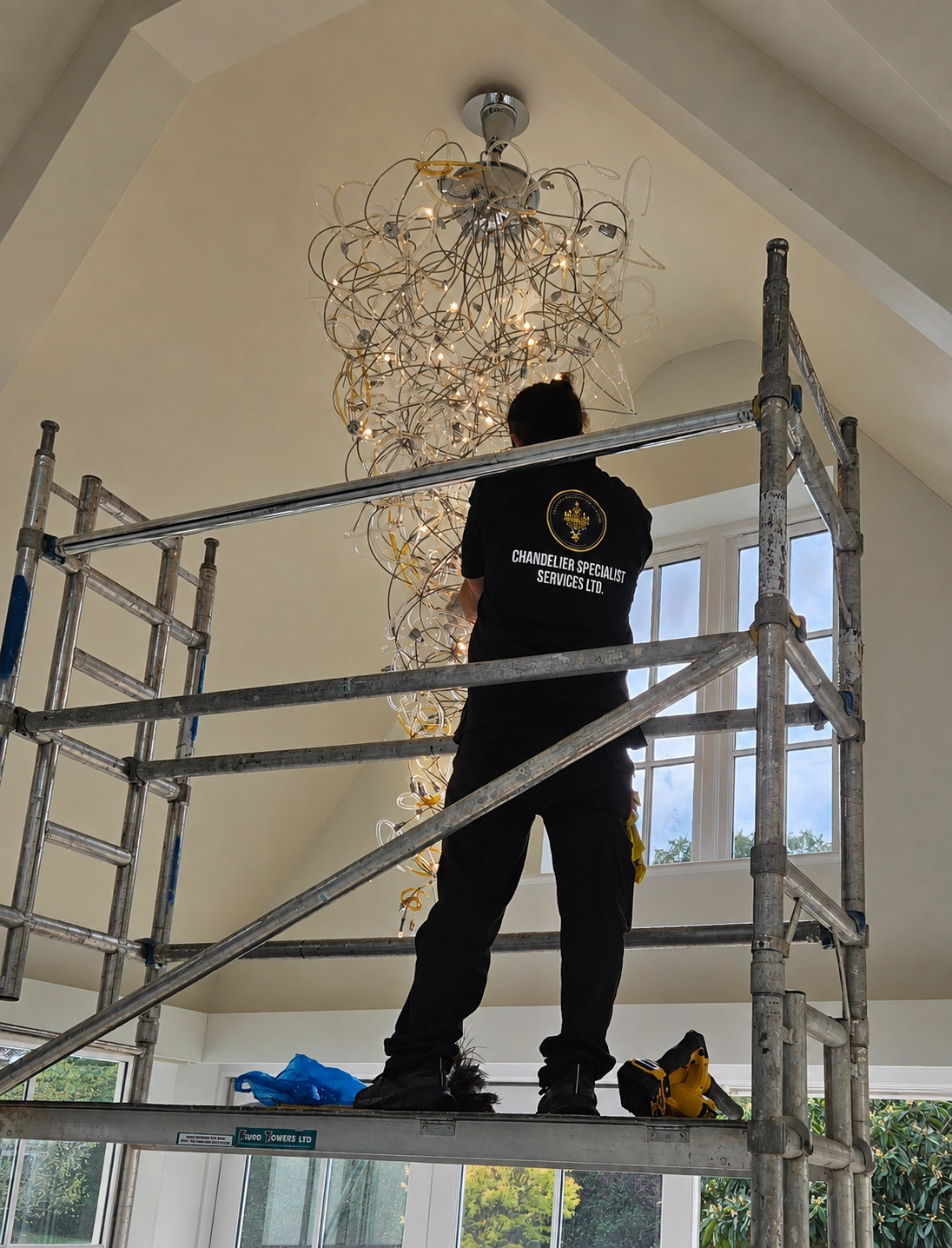 Chandelier specialist on a mobile scaffold tower servicing a looping wire and glass chandelier beneath a vaulted ceiling