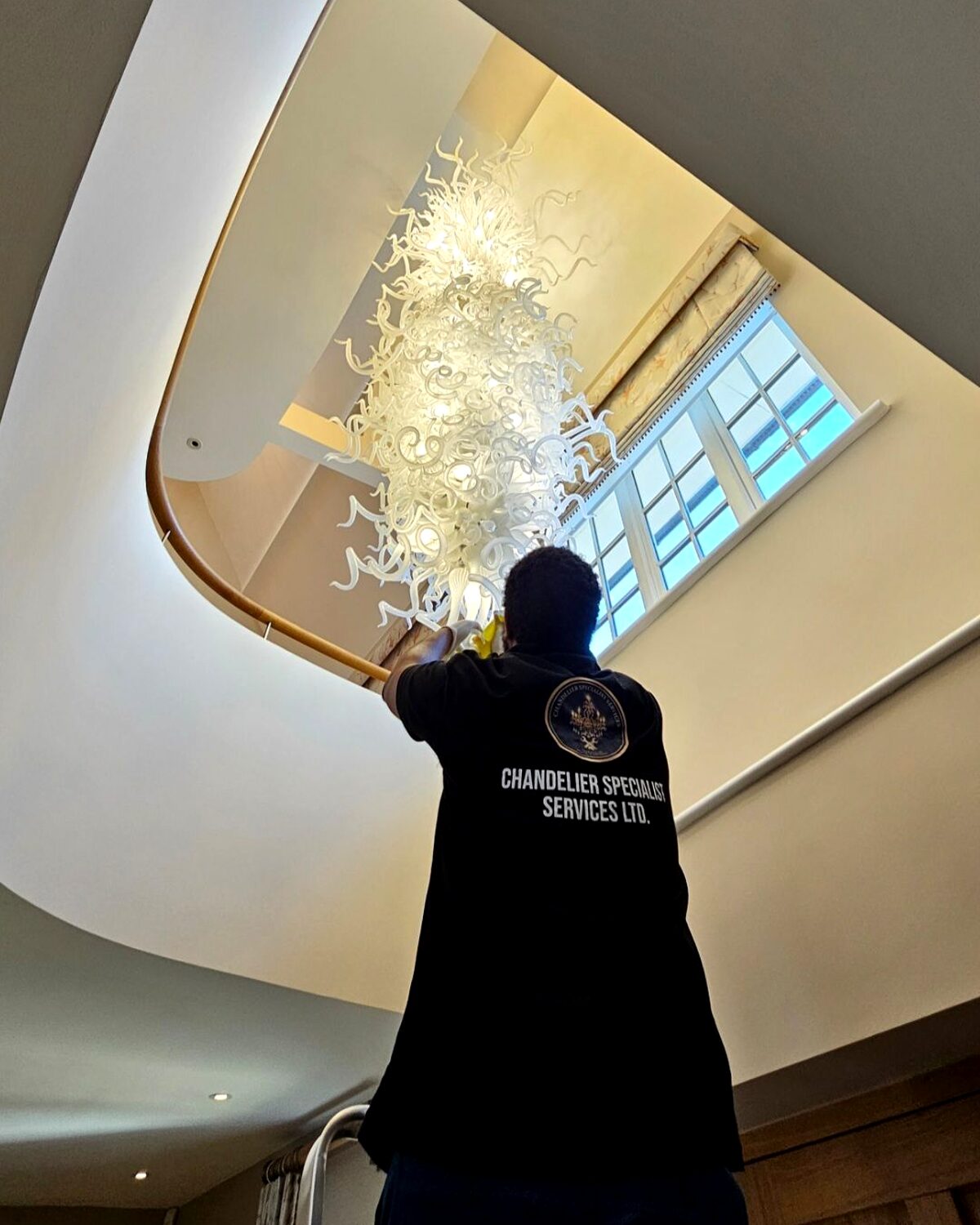 Chandelier specialist working beneath a white hand-blown glass swirl chandelier over a curved staircase