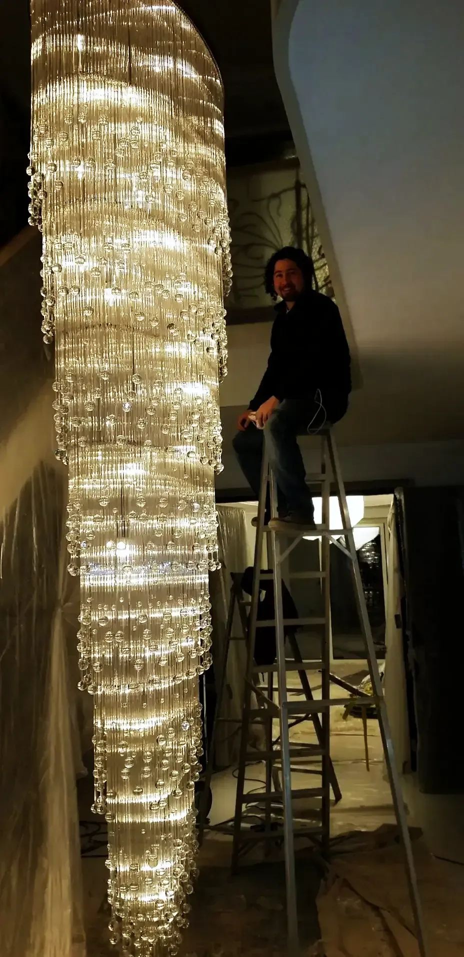 Specialist on a stepladder beside a tall crystal rod cascade chandelier during installation