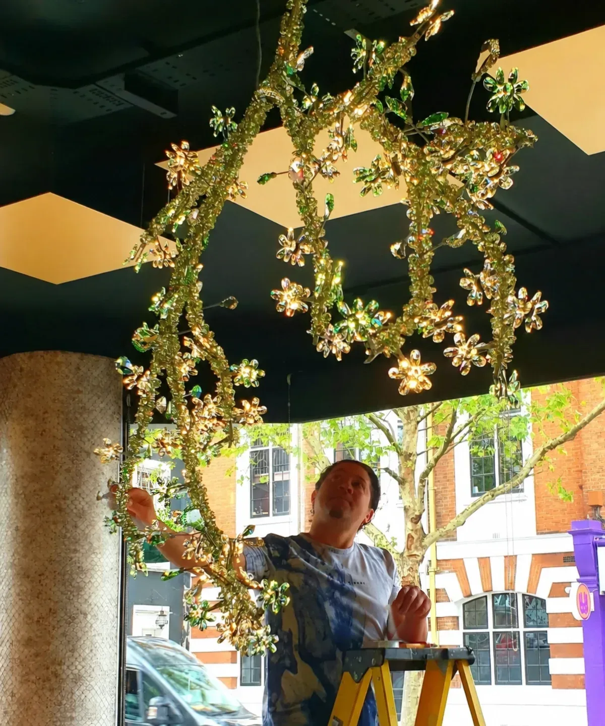 Specialist on a ladder adjusting a gold and green Swarovski crystal floral chandelier during installation