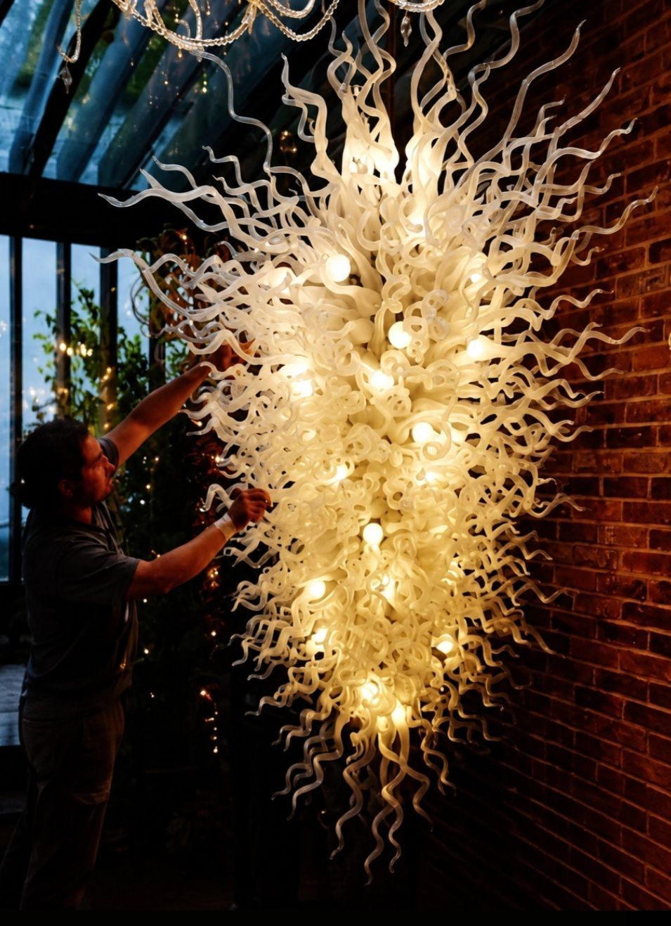 Chandelier specialist working beside a large white hand-blown glass chandelier against a brick wall